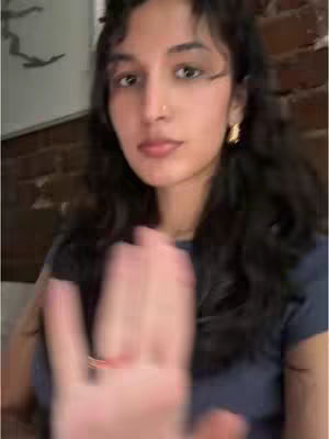 A young woman is centered in the frame, slightly blurred. She has long, dark hair, a small nose ring, and is looking at the camera. She's wearing a dark blue top and a gold earring. Her right hand is in the foreground, extending towards the camera, giving off a slight wave or stop gesture. The background is intentionally blurred, with hints of a brick wall.
