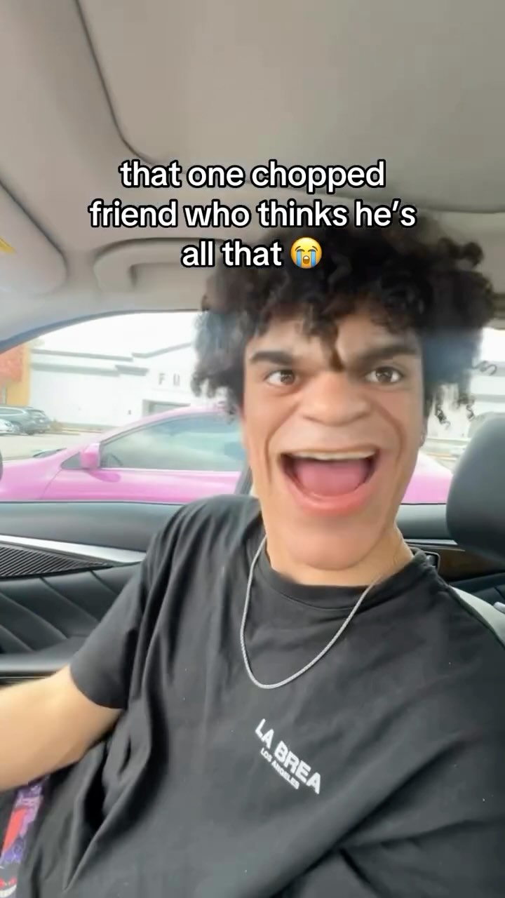 A selfie-style video shot inside a car featuring a young man. The man is in the foreground, making a wide-eyed, exaggerated expression with his mouth open. He's wearing a black t-shirt with white text, perhaps a brand name. Behind him, the interior of the car is visible, as well as a glimpse of a pink vehicle parked outside.