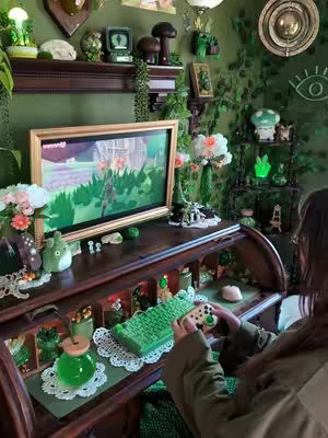 A cozy and detailed interior view. The foreground features a wooden desk filled with various green-themed decorations, including a green keyboard and controller, and a glass jar with a green liquid. In the background, a computer screen displays a video game on a desk with various collectibles and framed pictures. The walls are adorned with greenery, completing the immersive and thematic decor.