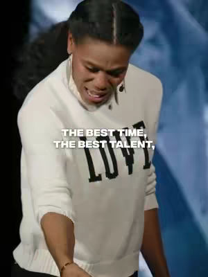 A medium shot of a woman in a white sweatshirt with the word "LOVE" in large black letters. She is speaking, and the image has a blurred background, possibly from a stage or studio setting. The overlay text adds context to her speech.