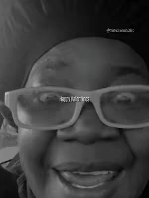 A grayscale image of a person's face in close-up, framed from the bottom up to the midline of the forehead. They wear thick-framed, light-colored glasses with the text 'Happy Valentines' superimposed across the lenses of the glasses, which reflect the image of the environment. The person's facial expression is jovial and slightly upturned, suggesting a smile. The lighting is even, emphasizing the details and a hint of background. The overall composition is simple, with a focus on the person's face and the message.