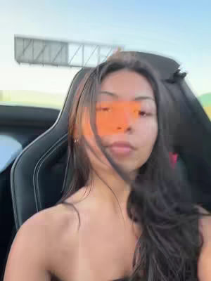 A young woman is seen from the chest up, looking towards the camera. She is in a car, with her hair flowing. The sky is visible in the background, providing a bright backdrop to the scene.