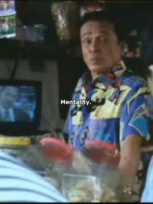 A man with short black hair and wearing a blue and yellow patterned shirt is looking towards the viewer. Behind him is a counter with items and a small TV showing two individuals talking. There is a text overlay that says in thin white font, 'Mentality.'