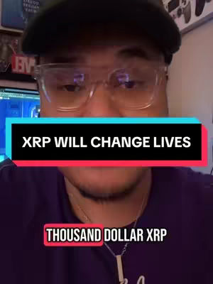 A selfie-style video cover image featuring a man wearing glasses and a baseball cap, partially obscuring his forehead. Overlaid on the image are text boxes with the phrases 'XRP WILL CHANGE LIVES' and 'THOUSAND DOLLAR XRP.' The background contains elements of a home setting, including what appears to be a computer screen.