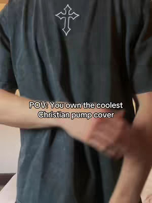 The image shows a person from the chest up, wearing a dark gray t-shirt with a white cross emblem on the upper chest. The person's arm is slightly raised, and the text overlay reads, 'POV: You own the coolest Christian pump cover'.