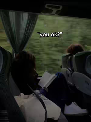 The image is taken from inside a bus or train. The main focus is on a person sitting by the window, reading a book. The person is wearing a cream-colored shirt and dark blue pants. The environment outside the window shows a blurred green landscape, suggesting the vehicle is in motion. Overlayed text reads "you ok?"