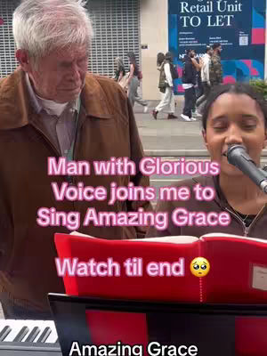 A woman sings into a microphone while an older man with white hair stands on the left, partly obscured, both in outdoor city settings with a sign in the background.