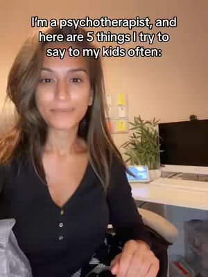 A woman with long brown hair is seated in front of a desk, looking at the camera. She is wearing a black top, with text overlayed.