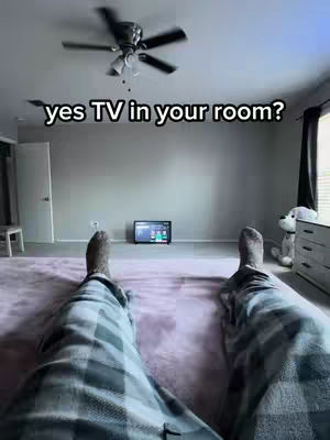 A first-person perspective shot from someone lying on a bed or a large rug. The feet, covered in a blanket or pajama bottoms with a plaid pattern are visible. In front is a small TV displaying a screen, likely reflecting content. The room has a gray wall, a white door, and a ceiling fan. The overall shot suggests a relaxing, casual atmosphere.
