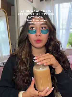 A woman is in the foreground, mid-shot, holding a large glass jar with a beverage in it. She has under-eye patches on and her hair is long, wavy and auburn. Text overlays the top of her head, and the background consists of a room with a mirror, a window, and cream-colored walls.