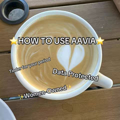 A close-up shot of a cup of latte art with a heart shape in the foam. The image is set on a wooden surface. Text overlays include "HOW TO USE AAVIA" with star emojis, "Tinder for your period," "Data Protected," and "Women-Owned."