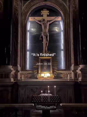 The image shows a dimly lit church interior, focusing on a religious display. At the center is a crucifix with the figure of Jesus Christ. Below the crucifix is a golden frame with an image, and a small display of lit candles in the foreground. The words "It is finished" and the verse "John 19:30" are added as text overlay on the image.