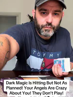 A man with a beard is the central focus, wearing a dark blue t-shirt that says 'TOMMY' and a gray hat. The image is lit by a bright interior light, with the man holding a deck of cards in his right hand and a collection of objects and items laid out in front of him.