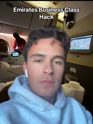 A young man with short dark hair wearing a light blue hoodie is the central focus, set inside an Emirates business class cabin. Behind him, a person in a red shirt is visible. The video's title, 'Emirates Business Class Hack,' is displayed in a white font at the top. The cabin features a monitor on the right and various interior details with soft, warm lighting.