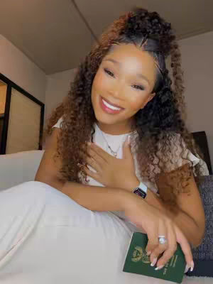A woman with curly hair, wearing a white top, holding a green passport and smiling. Her hand is touching her chest, and she is seated in a well-lit room.