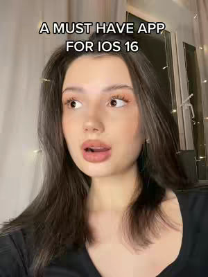 A woman with long dark hair is looking slightly off-camera with a surprised expression. Above her head, in a black font, are the words 'A MUST HAVE APP FOR IOS 16". The background is blurred, with hints of a window and string lights.