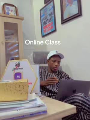 A young man wearing a white cap sits at a wooden desk with a laptop, suggesting an online class or tutorial. The background features a framed poster reading 'Forget the mistake, Remember the lesson.' A framed picture is on the right side. A desk displays books and papers. The lighting suggests an indoor setting.