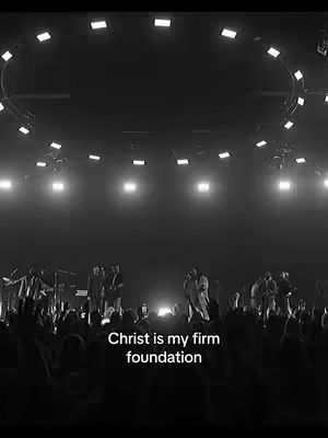 The image is a black and white shot of a live event or concert setting. It features a stage with performers at the bottom, their silhouettes visible against stage lighting. In the background, there's an audience, also silhouetted, with their hands raised. The ceiling has what appears to be a series of overhead lights arranged in a semi-circle, adding to the ambiance.