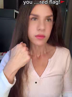 A woman with long brunette hair is shown from the chest up, slightly angled towards the camera. She is wearing a light pink, ribbed henley shirt. In the background, there is a glimpse of a dark cabinet or appliance. Text is overlaid at the top of the frame.