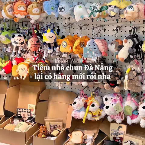 The cover image showcases a display of various plush keychains and stuffed animals hanging on a grid. Below the display are cardboard boxes filled with what appears to be packaged goods and gifts. Text overlays, including the words "Tiệm nhà chun Đà Nẵng" and additional Vietnamese text, are visible. The overall composition suggests a store or online shop showcasing its merchandise.
