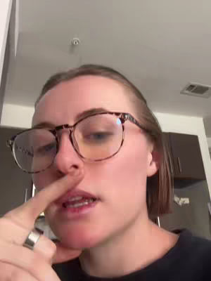 A close-up shot of a person pointing their index finger at their nose. The individual wears glasses, a dark t-shirt and has a ring on their finger, with a neutral background.