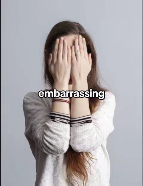 A young woman, seen from the chest up, has her hands covering her face. The fingers are spread, partially obscuring her features. She wears a knitted white sweater against a simple gray background. Text in a bold font is superimposed across the image.
