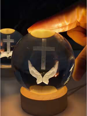 A clear glass sphere with a cross and hands image inside, sitting on a wooden base, with a hand of the presenter. The lower portion of the sphere has a hand-shaped image with a light-up effect. The wooden base has a yellow light-up effect as well.