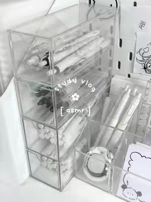 A transparent acrylic desk organizer filled with office supplies. The organizer has multiple compartments and drawers, filled with white pens and other stationery. The overall aesthetic is clean and minimalist.