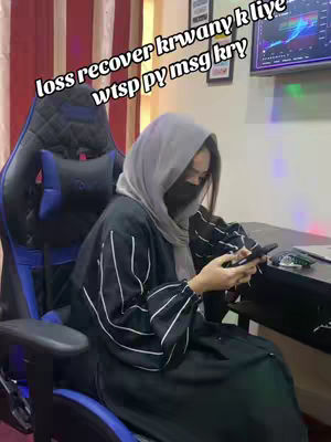 A woman wearing a gray hijab and a black face mask is seated in a gaming chair. She is holding a smartphone and looking at the screen. The background includes a desk, a wall with pictures, and a computer screen. Text overlays the image in a white font.