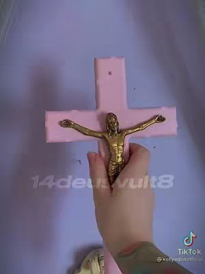 A person's hand holds a pink cross featuring the golden figure of Jesus. The hand, with a tattoo on the forearm, is in the foreground against a blurred background of a light purple wall.