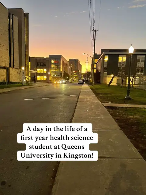 The image captures an evening street scene at Queens University in Kingston. The foreground shows a sidewalk and road with cars. Buildings with lit windows line the sides. The sky transitions from blue to orange at the horizon. Text overlays on the image.