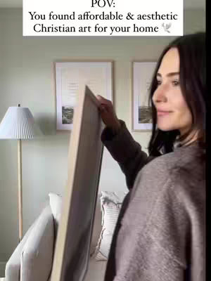 A woman holding an artwork in front of a wall with framed pictures, a lamp, and a sofa.