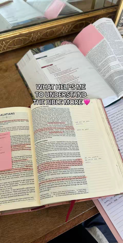 The image shows an open Bible with notes and highlighted text, alongside other books and a pink sticky note. The background includes a reflective surface, suggesting a tabletop setting. The scene is well-lit and organized, giving a sense of study and reflection.