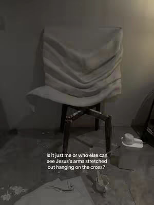 The image shows a chair draped with a white cloth in a dimly lit room. The cloth's folds resemble a figure with outstretched arms, evoking the image of Jesus on the cross. A few objects are in the foreground.