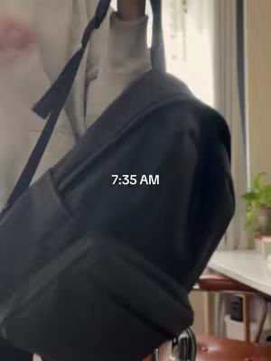 A person is holding a black backpack with the strap over their shoulder. The time 7:35 AM is overlaid on the image. The background consists of a window and a hint of a green plant.
