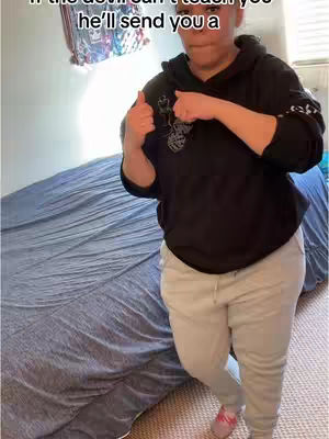 A person in a black hoodie and gray sweatpants is standing in a bedroom, giving two thumbs up. The background includes a bed with a blue comforter, a patterned wall hanging and a window.