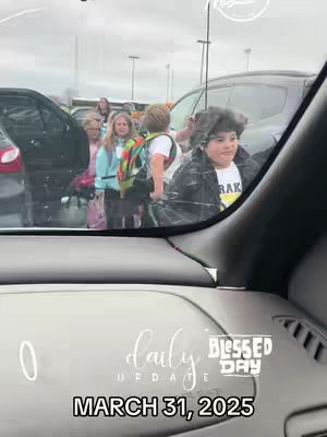 The image captures a scene from inside a car, looking out at a group of children exiting a building, presumably a school. The focus appears to be on a young boy wearing a jacket, with several other children in the background. The shot is taken through the windshield, and overlaid text indicates a daily update along with the date.