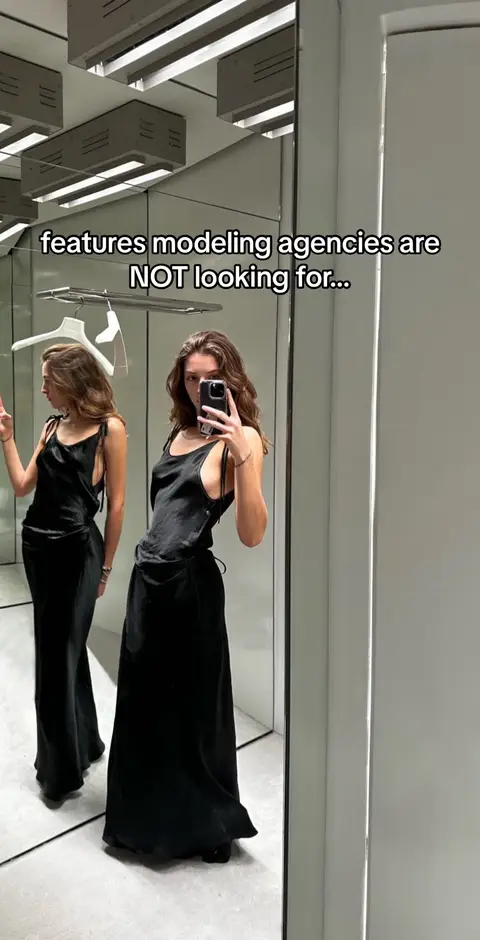 A woman in a black slip dress is standing in a room-length mirror, taking a selfie with her phone. The room has a bright ceiling with long, rectangular lights. The woman is positioned toward the right of the frame, angled slightly, and her reflection is seen behind her. Text on the image reads, features modeling agencies are NOT looking for...