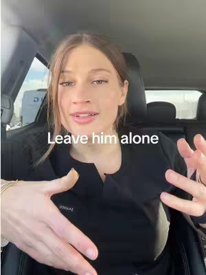 A woman with long brown hair is sitting in the driver's seat of a car, looking directly at the camera and speaking. She has fair skin and is wearing a black top. The background blurs to reveal a possible outdoor setting.