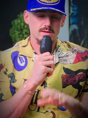 A man in a blue cap and yellow shirt with a microphone in hand, ready to speak into it. The background is blurred, with hints of green foliage.