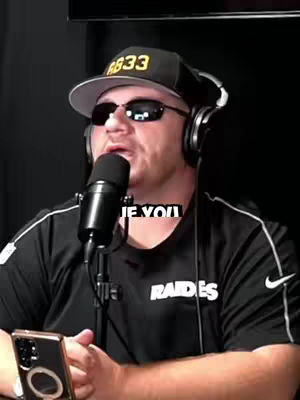 A medium shot of a man with dark sunglasses and a black cap, headphones, and a microphone, set against a black background. The man is wearing a black Raiders jersey with the team name in white. Text overlay in the middle says "IE YOU". A gold-colored phone is in the person's hand on the left side.