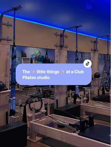 A pilates studio interior shot with equipment in the foreground and background. The room is well-lit with a blue ceiling and warm-toned walls. Text overlay is added.