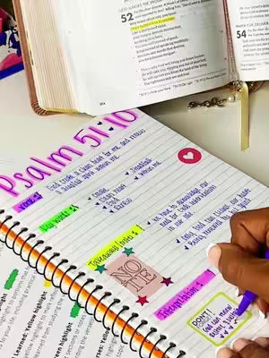 The image shows a hand writing notes on a spiral notebook, with a bible open in the background. The notebook has colorful highlights and notes. The background has a table where a bible is open and a book in a brown cover.