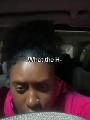 A close-up shot of a person's face inside a car. They have short, dark, curly hair. The person is looking down with a slightly surprised or concerned expression. The background includes the car's interior.