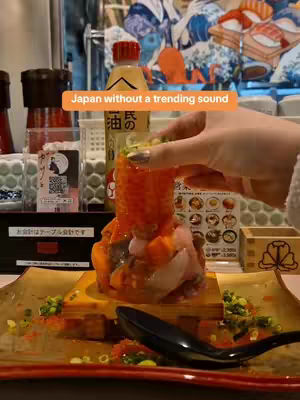 A close-up shot shows a beautifully arranged sushi dish on a golden plate. The sushi is stacked high with layers of textures and colors, including bright orange roe, various cuts of fish, and garnishes. Behind the dish are elements of a restaurant setting, like bottles and menu items.