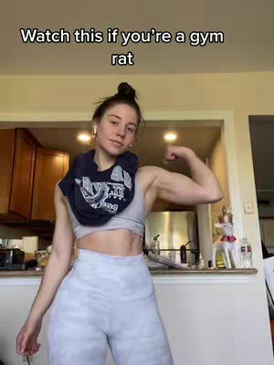 A woman flexes her bicep in a kitchen setting. She is wearing a gray sports bra, gray leggings, and a dark blue shirt draped around her neck. Text overlay reads, 'Watch this if you're a gym rat'. The background shows a kitchen with cabinets and a counter.
