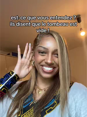 A woman with blonde-highlighted hair, wearing a white top with a blue embroidered design, smiles and gestures towards her ear with her hand. Overlayed text in black informs the viewer.