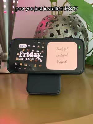 An iPhone is displayed on a wireless charger, showing the Standby mode on its lock screen. The screen displays the time, date, and a custom widget with the words "thankful, grateful, blessed."