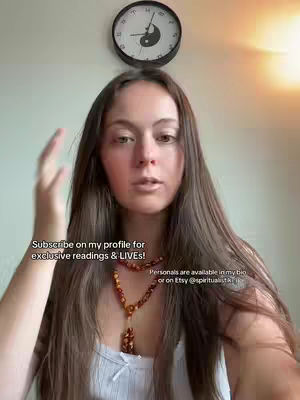 A medium shot shows a woman with long brown hair looking directly at the camera. She wears a white top and a necklace. Behind her is a wall with a clock and a light source creating a soft glow.