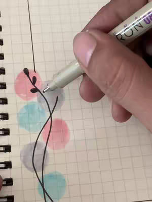 A hand holding a pen is drawing vines and leaves on top of colorful watercolor circles inside a spiral notebook. The image is a close-up shot, only showing the drawing hand and notebook.